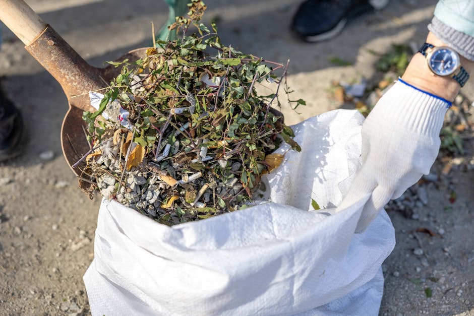A person wearing a light-colored work glove is holding the edge of a large white waste bag filled with garden debris, including green leafy branches, small twigs, and crushed stones or gravel. The bag